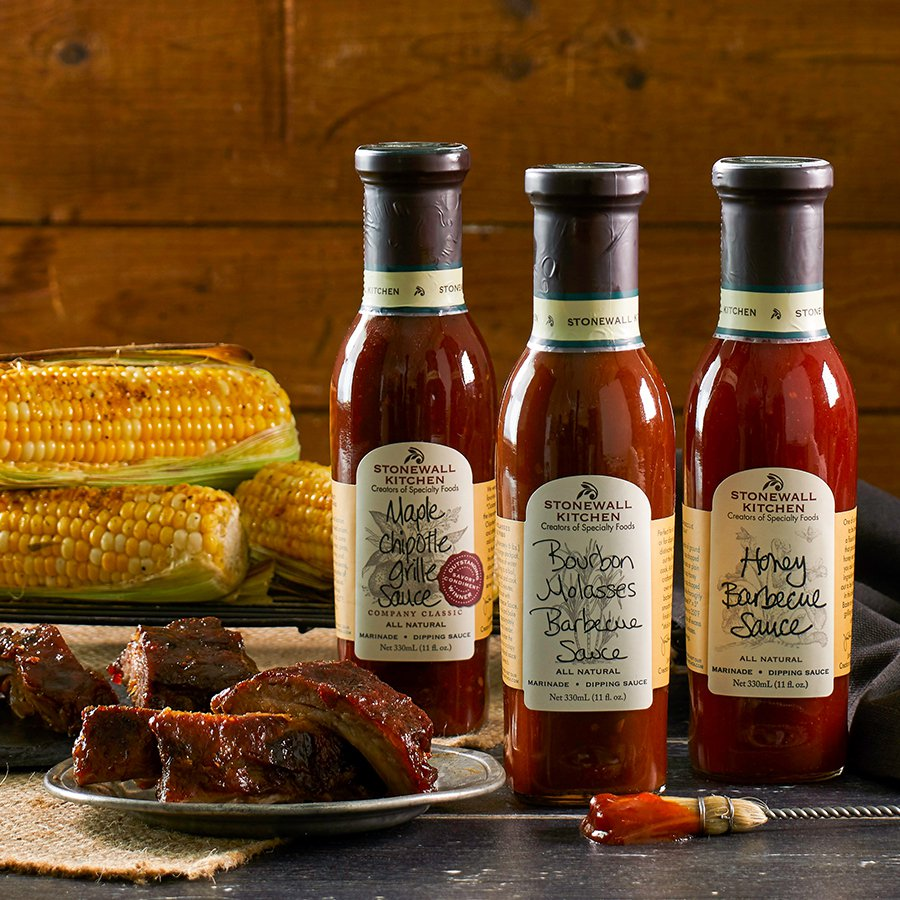 Drei Flaschen Stonewall Kitchen Barbecue-Saucen neben Grillgerichten auf Holztisch