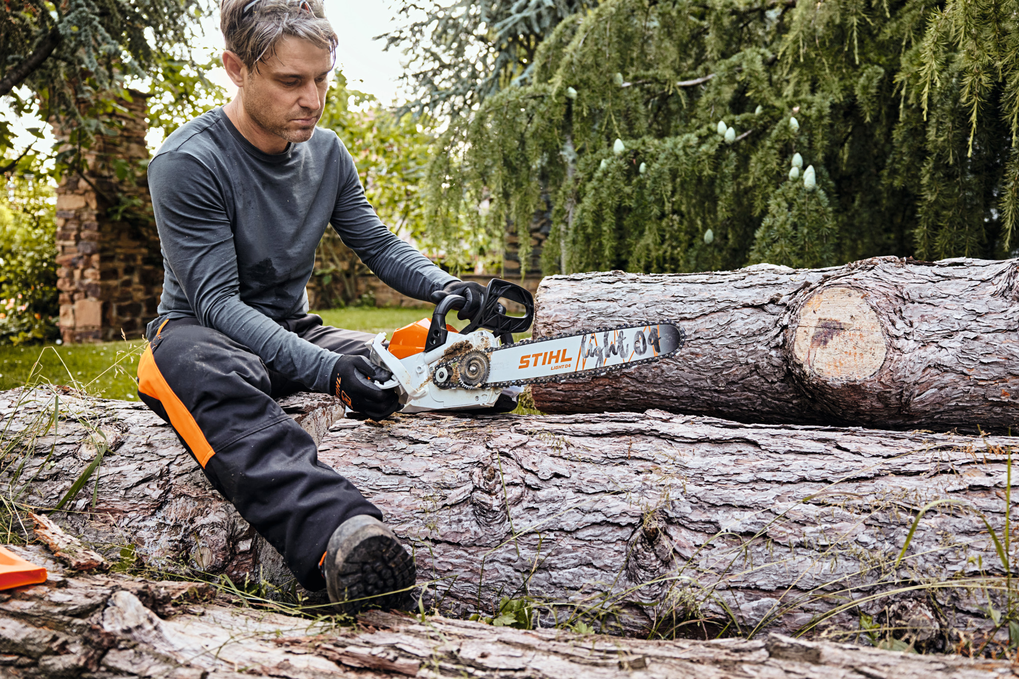 Mann überprüft STIHL Akku-Kettensäge auf Baumstamm in grünem Garten