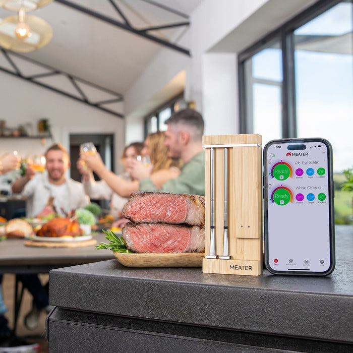 Smartes Fleischthermometer MEATER Pro Duo neben Smartphone mit App und feiernde Personen im Hintergrund