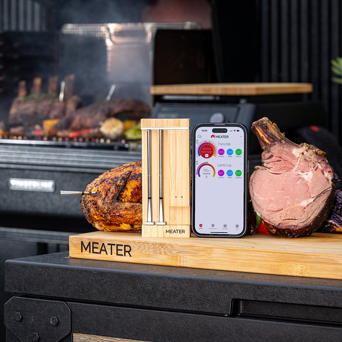 Smartes Fleischthermometer MEATER Pro Duo neben gegrilltem Hähnchen und Lammkeule auf Holzbrett mit Smartphone-App