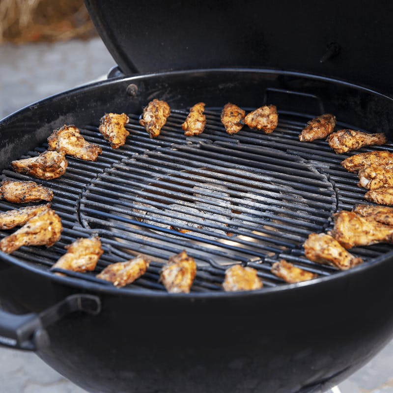 Geöffneter schwarzer Kugelgrill mit knusprig gegrillten Hähnchenflügeln auf einem Grillrost