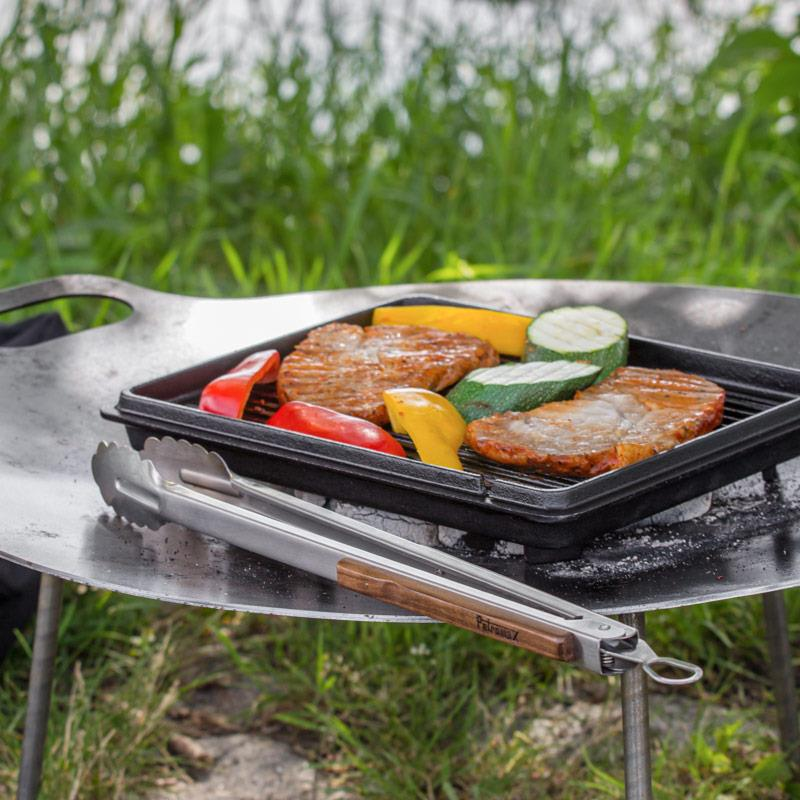 Petromax Grill- und Kohlenzange auf Grillplatte mit Fleisch und Gemüse im Freien