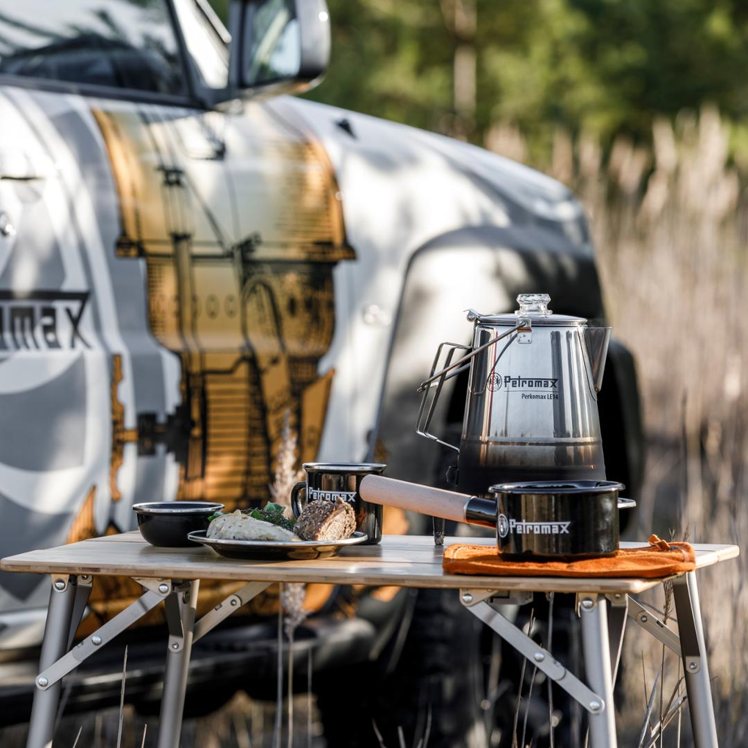 Outdoor-Kochszene mit Petromax Emaille Stieltopf und weiterem Kochgeschirr auf einem Campingtisch