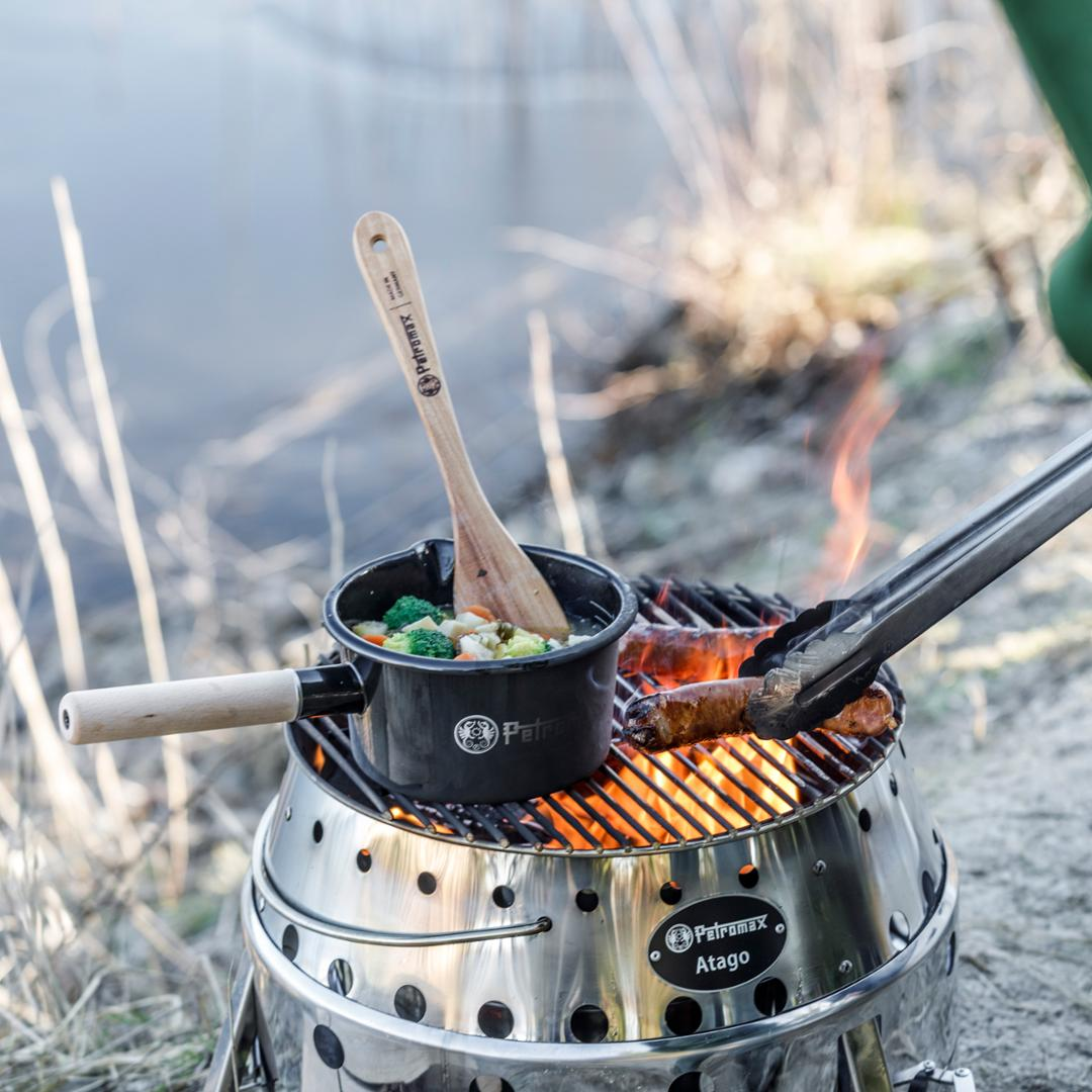 Petromax Emaille Stieltopf auf Outdoor-Grill am Seeufer mit Gemüse und Wurst über Feuer