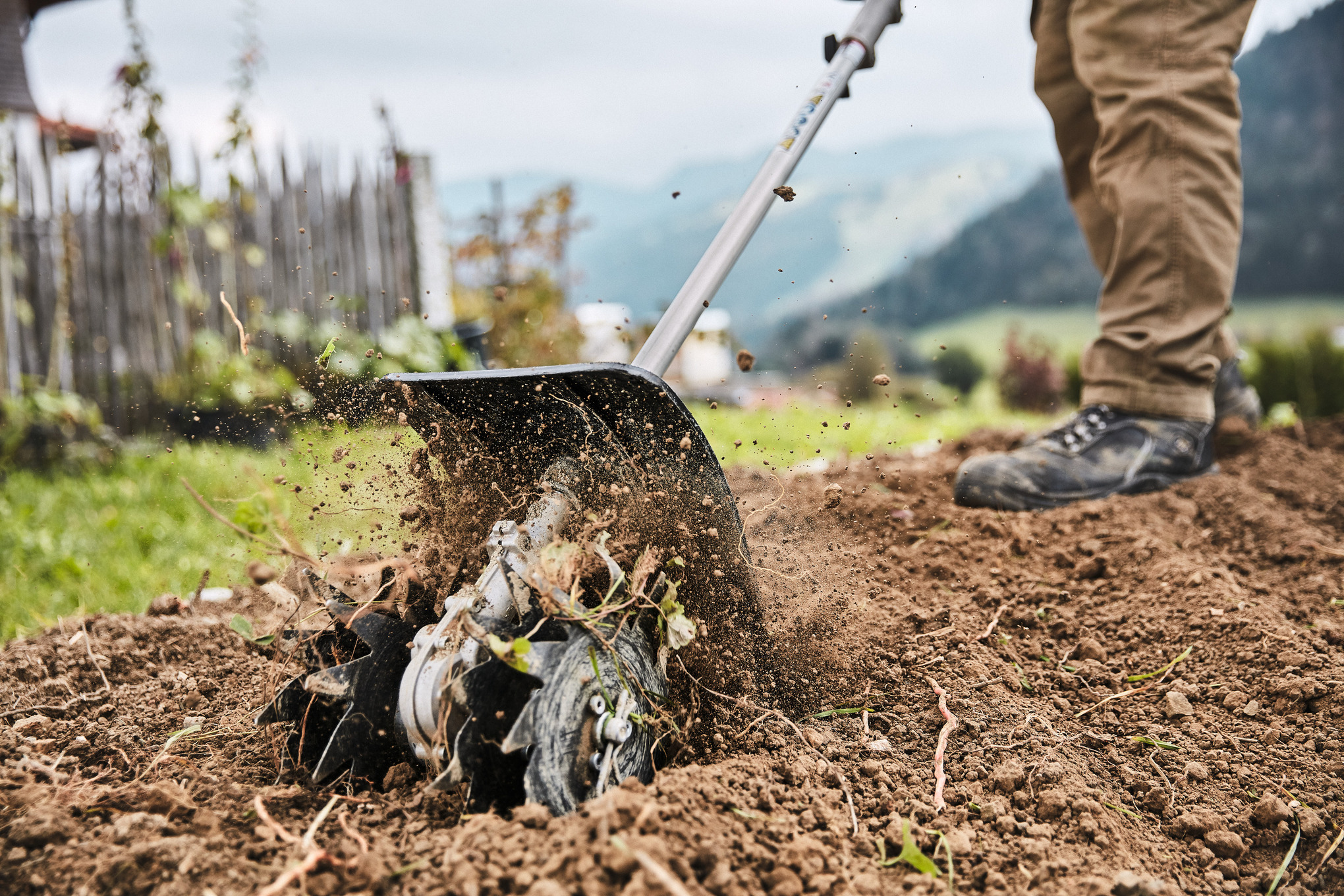 Person verwendet Stihl Bodenfräse in einem Garten mit aufgewirbelter Erde und ländlichem Hintergrund