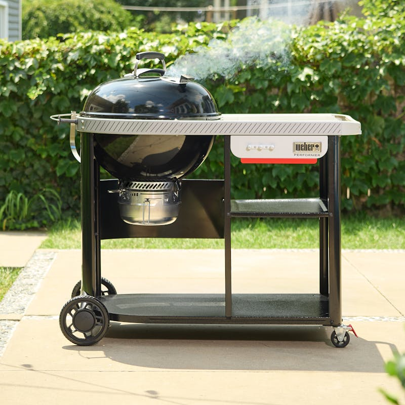 Weber Performer Premium Holzkohlegrill auf Rollwagen mit Rauch und Gartenhintergrund