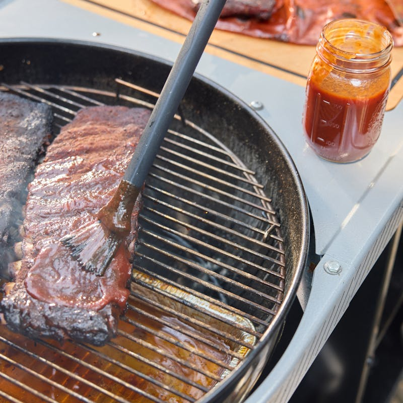 Weber Performer Premium Holzkohlegrill mit Fleischstücken und roter Sauce auf seitlichem Tisch