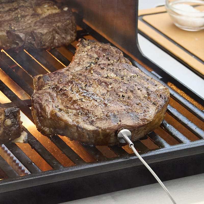 Steak mit Temperaturfühler auf einem Gasgrill, nebenbei eine Schüssel mit Sauce