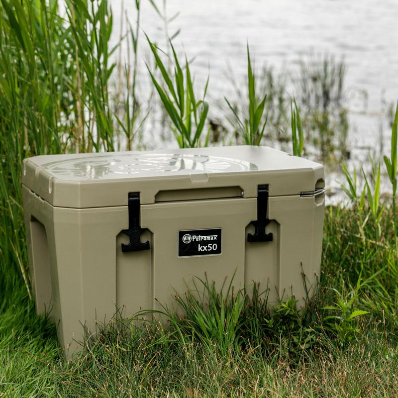 Olivgrüne Petromax Kühlbox kx50 am Ufer eines Gewässers umgeben von Gras