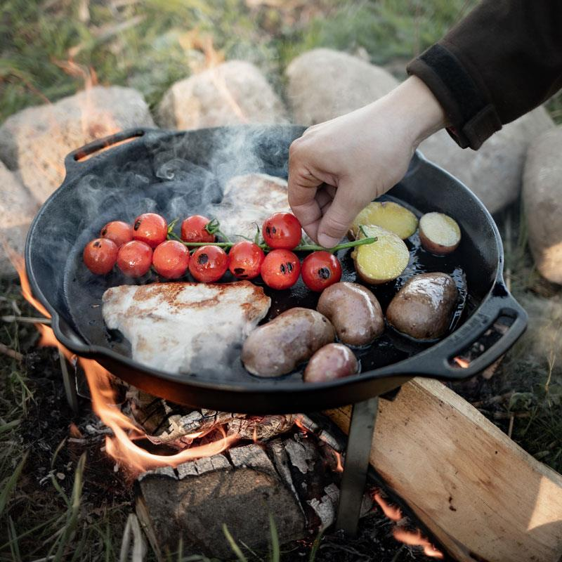 Person kocht Fleisch, Kartoffeln und Tomaten in gusseiserner Pfanne über Lagerfeuer