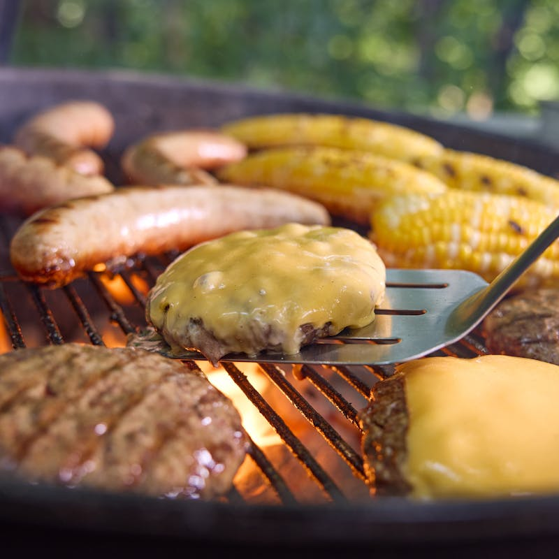 Burger und Würste auf einem Holzkohlegrill mit Maiskolben im Hintergrund