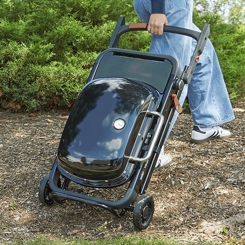 Person zieht zusammengeklappten Weber Traveler Compact Gasgrill auf Rädern im Freien