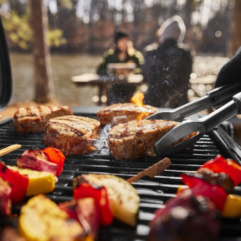 Steaks und Gemüse auf Weber Traveler Compact Gasgrill in natürlicher Umgebung mit Personen im Hintergrund