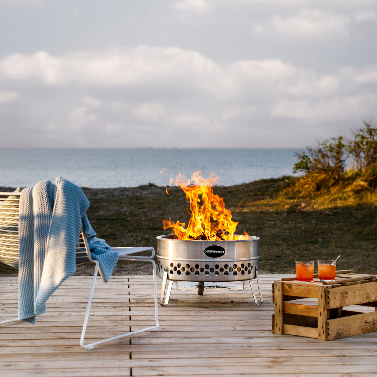 Petromax Feuerschale Tyropit auf Holzterrasse am Strand mit lebhaftem Feuer und Meerblick