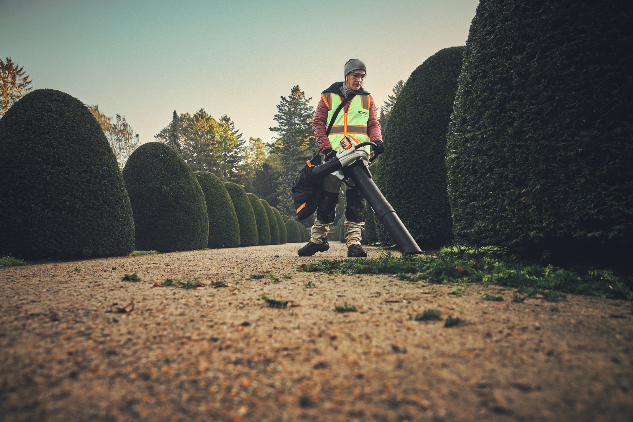 Professioneller Gärtner verwendet STIHL Akku-Saughäcksler auf Kiesweg neben hohen Hecken