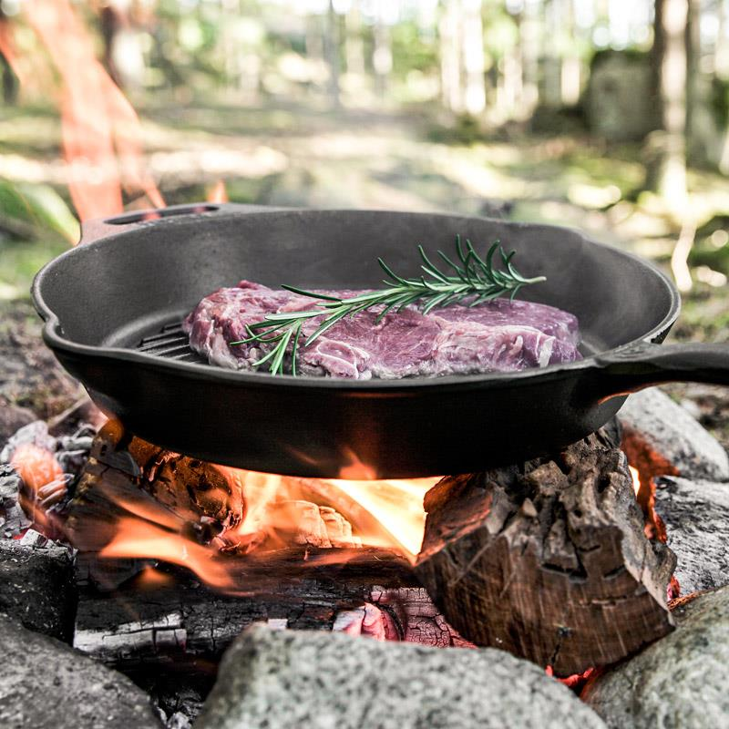 Petromax Gusseisenpfanne auf offenem Feuer im Wald mit bratendem Fleisch und Rosmarin