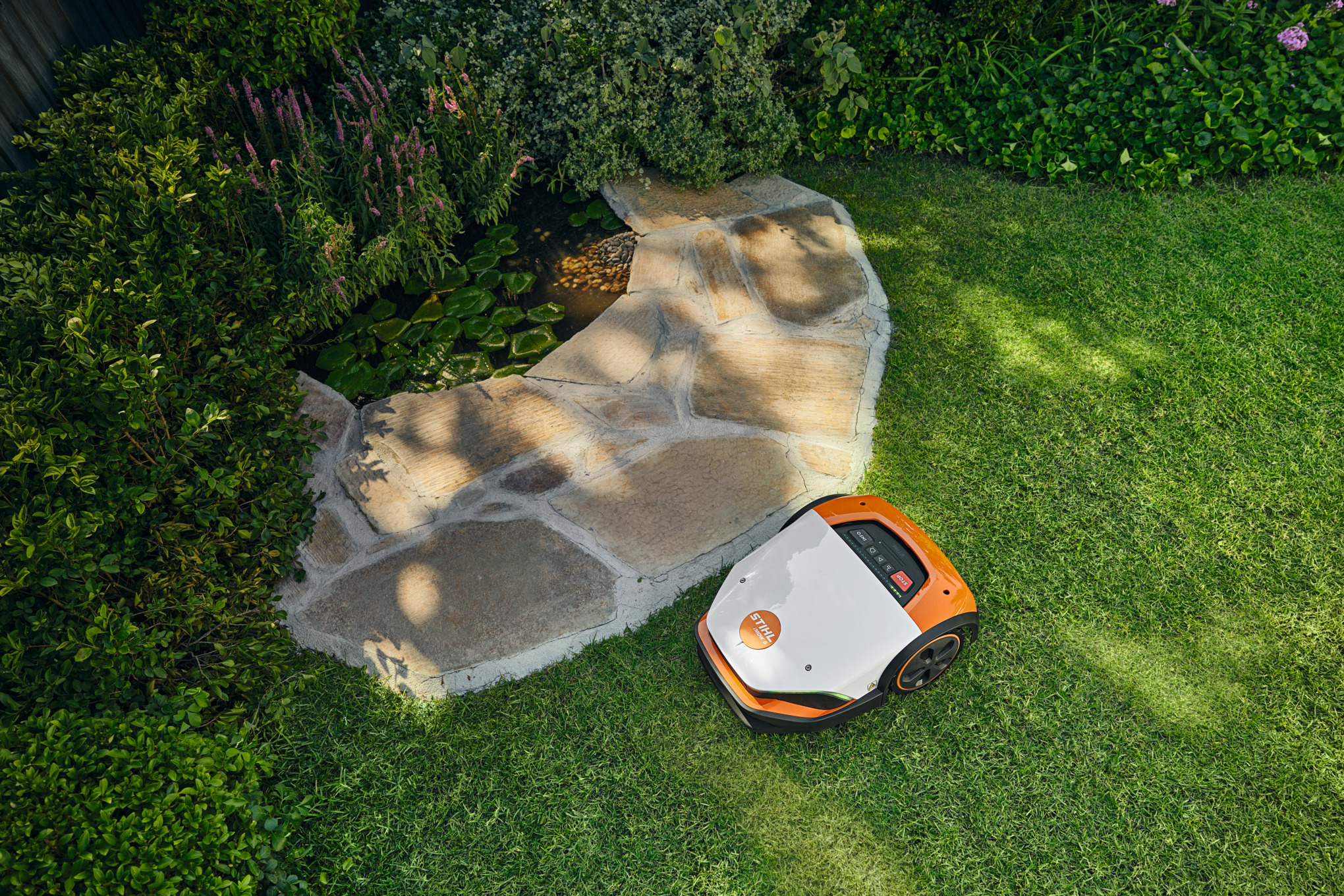 STIHL iMOW 7 Mähroboter mäht selbstständig einen grünen Rasen umgeben von Blumen und einem Teich