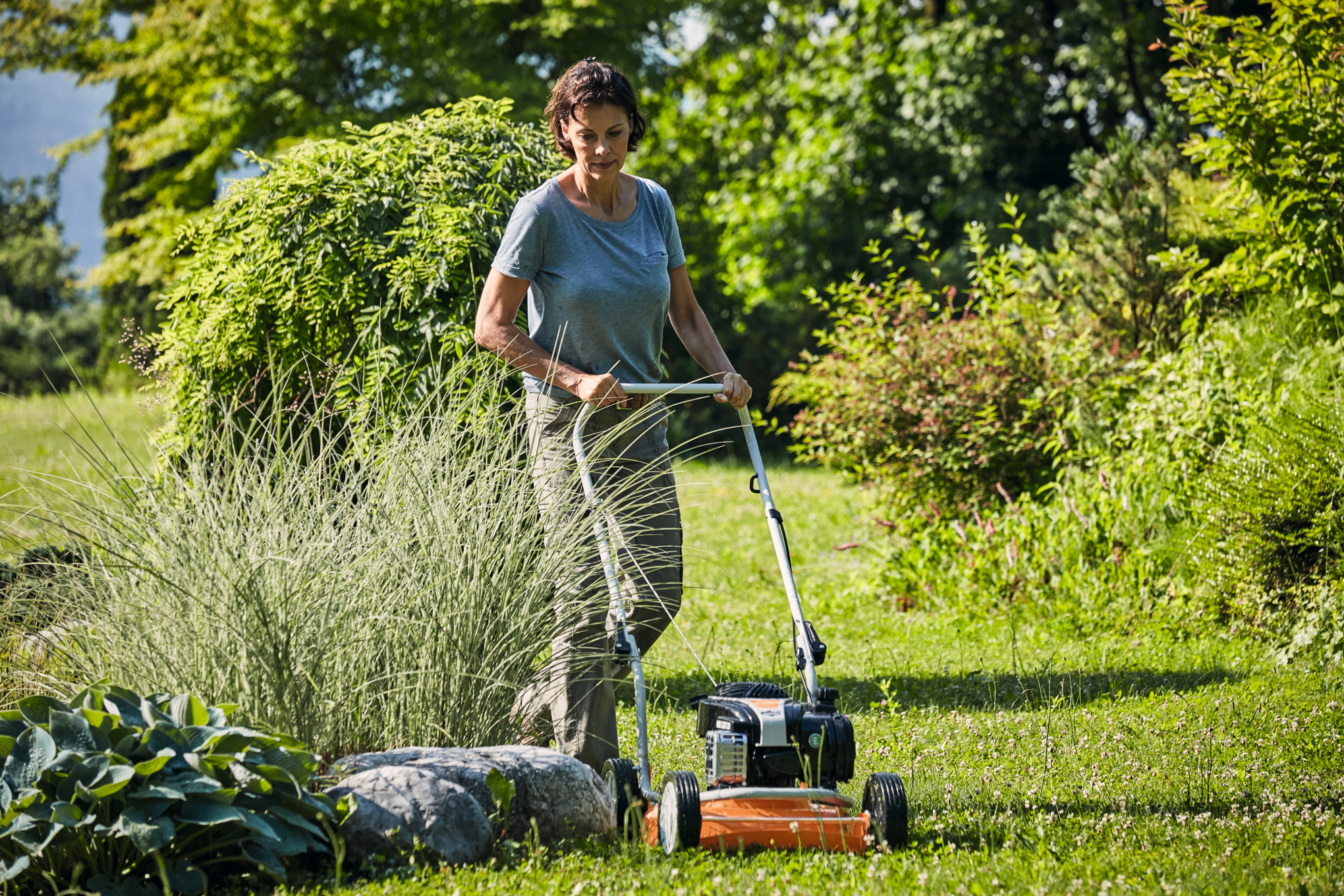 Frau mittleren Alters mäht Rasen mit STIHL RM 2 R Rasenmäher in einem sonnigen Garten