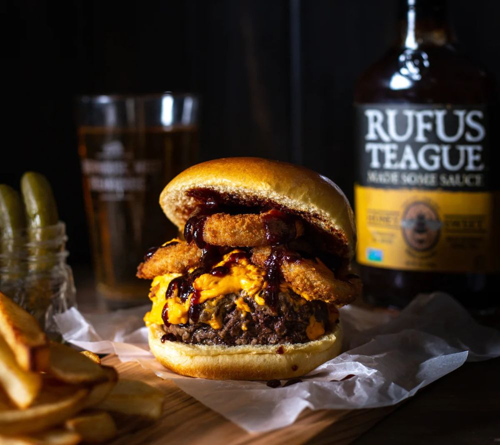 Burger mit BBQ-Sauce, Bier und Gurken im Hintergrund eine Sauce-Flasche