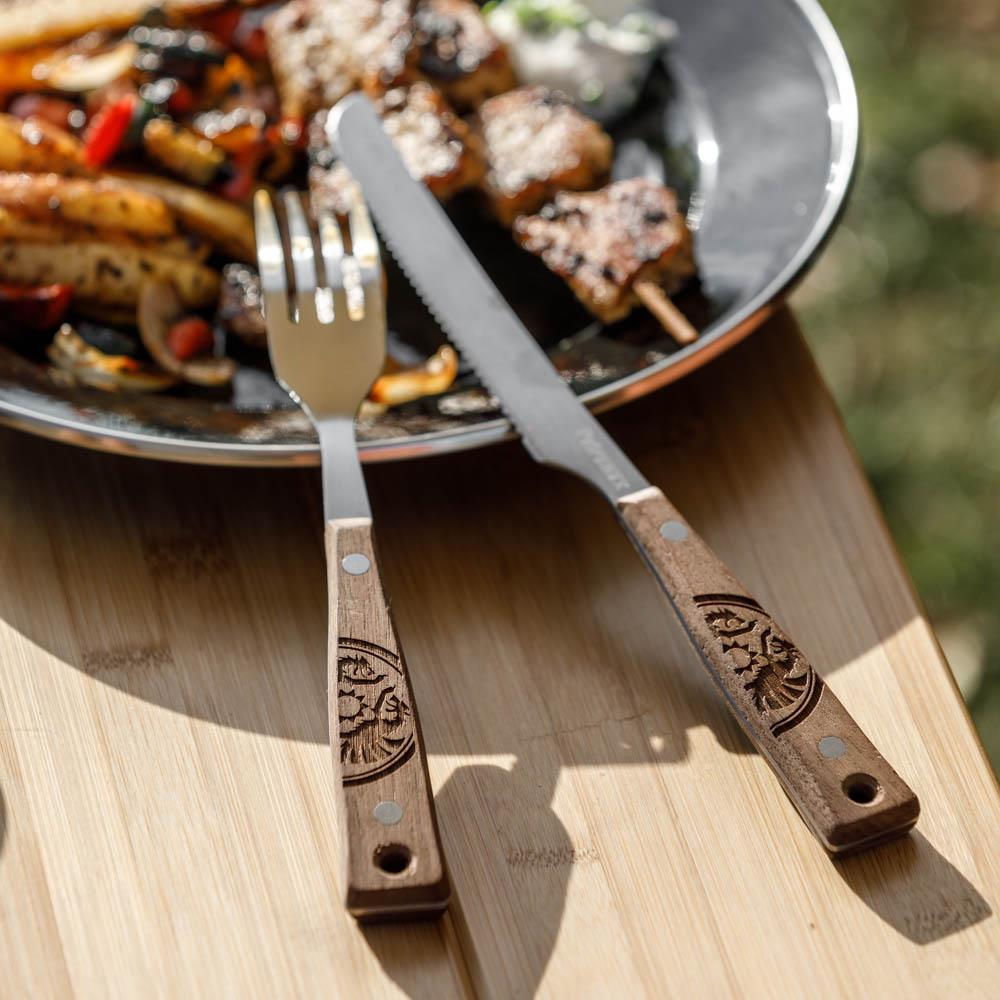Petromax Camping Besteck auf Holztisch mit gegrillten Speisen im Hintergrund