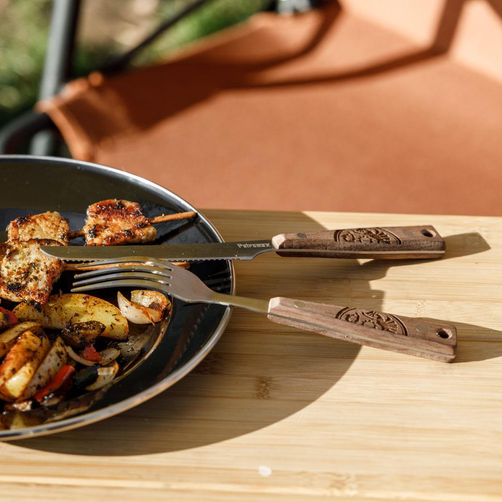 Petromax Camping Besteck auf Holztisch neben Teller mit gegrilltem Essen