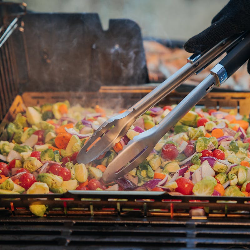 Weber CRAFTED Grillkorb mit Gemüse auf einem Grill, Person wendet Gemüse mit Zange