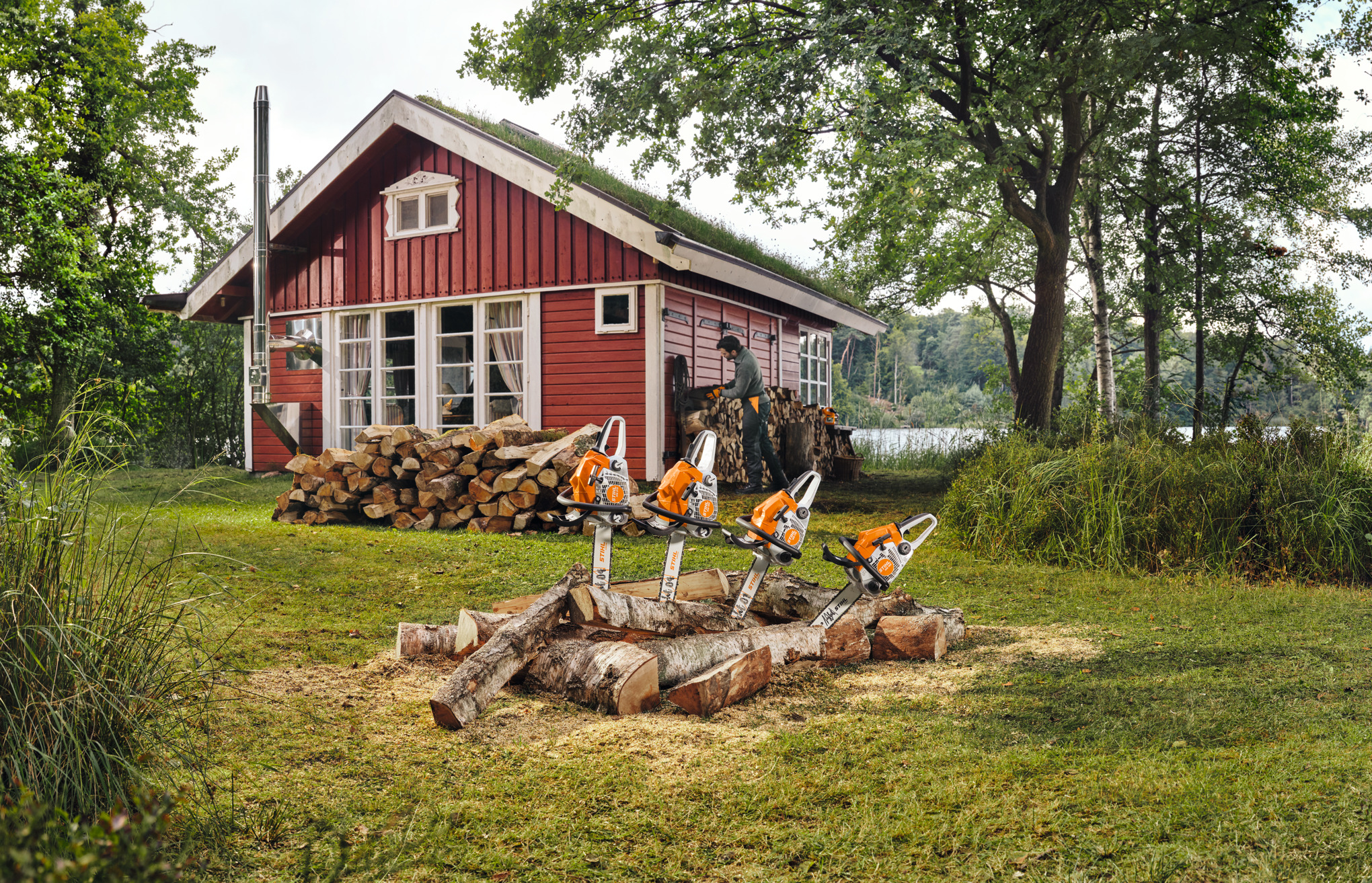 Rote Holzhütte mit Stihl Kettensägen auf Baumstämmen und Mann beim Holzstapeln