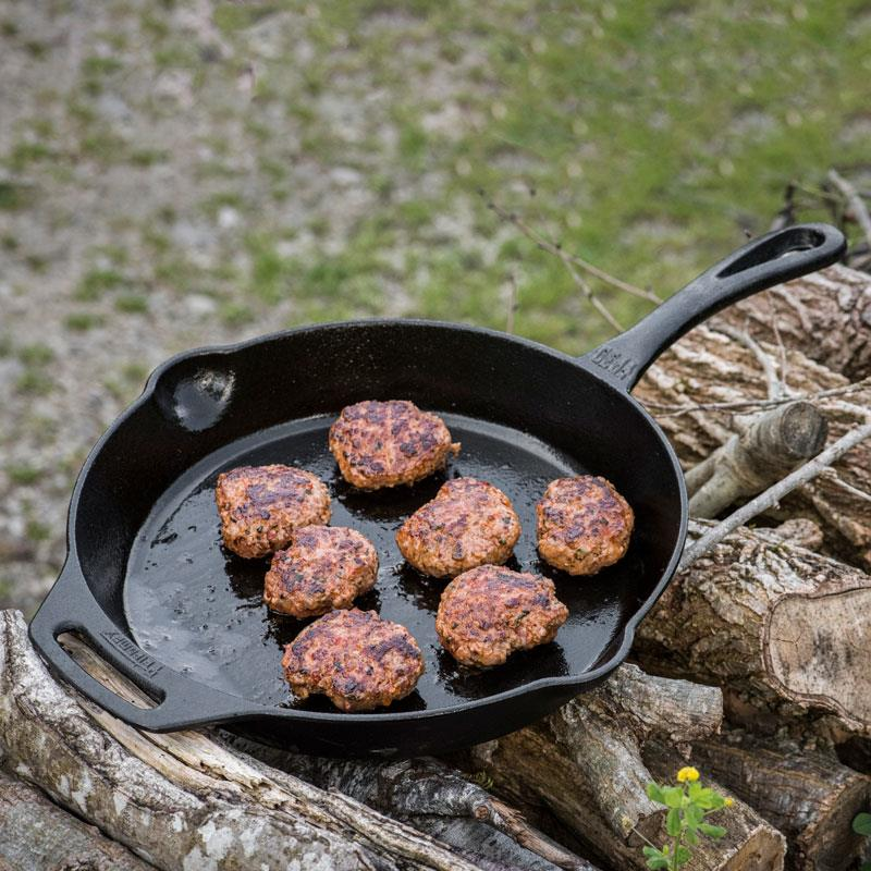Gusseiserne Petromax Feuerpfanne mit Fleischküchlein im Freien auf Holzhaufen