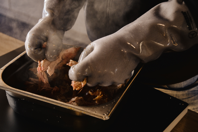 Person verwendet graue Silikon-Grillhandschuhe beim Zerteilen von Fleisch in der Küche