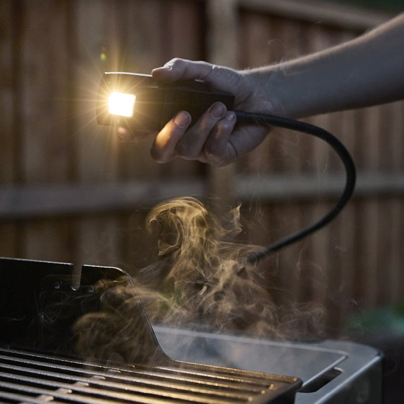 Hand hält eine flexible LED-Grilllampe über einem rauchenden Grill bei Dämmerung