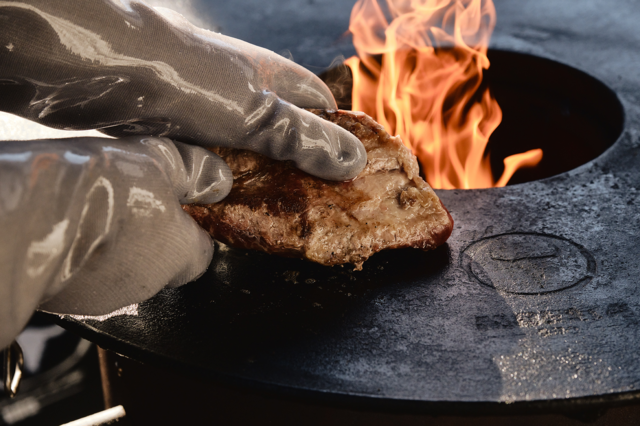 Hand in hitzebeständigem Silikonhandschuh wendet Fleisch auf Grillplatte neben offenen Flammen