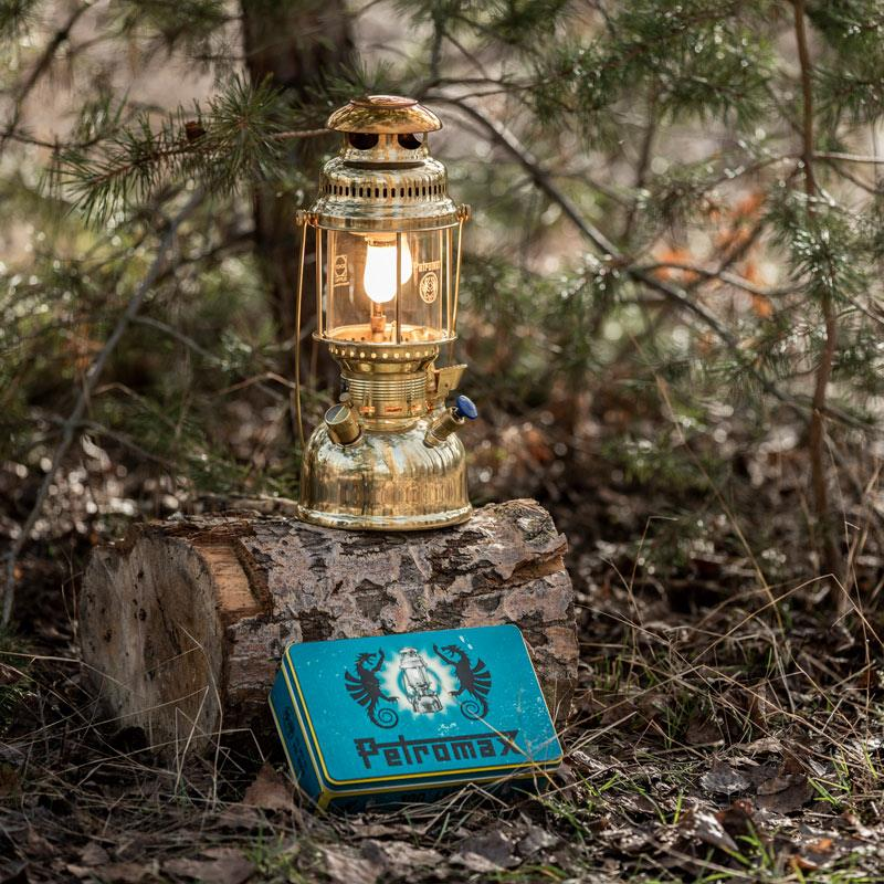 Petromax HK500 Petroleumlampe in einem Wald auf einem Baumstumpf neben einer blauen Logo-Box
