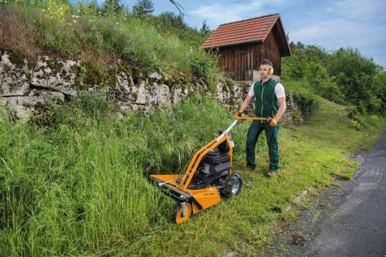 Mann bedient AS 63 2T ES Rasenmäher auf steilem Grashang neben Steinwall und Holzhütte
