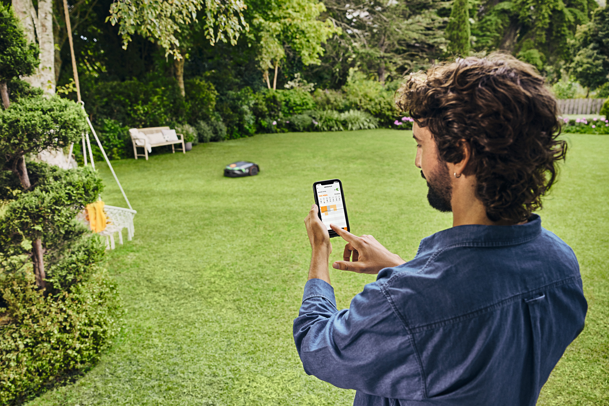Mann bedient Smartphone im Garten während Mähroboter Rasen mäht