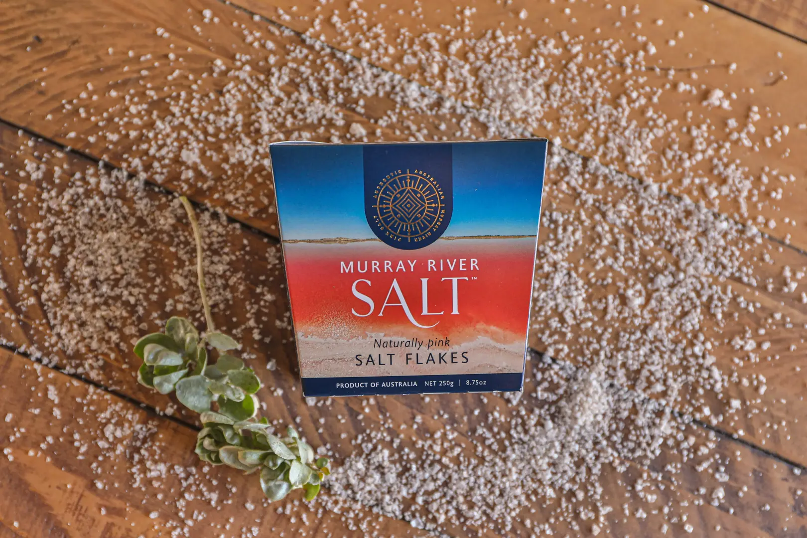 Packung Murray River Salt auf Holzuntergrund mit verstreuten Salzflocken und kleiner Pflanze