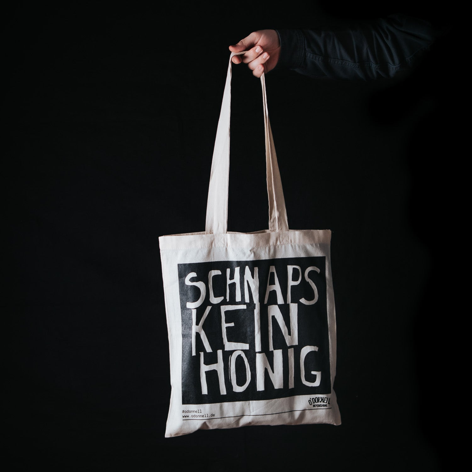 Person hält Jutebeutel mit Aufschrift 'Schnaps, kein Honig' vor schwarzem Hintergrund