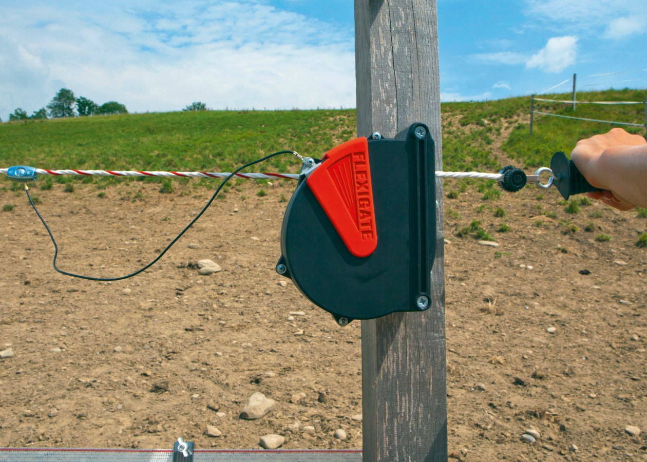 Flexigate Seilanschlusskabel an Holzpfosten auf Pferdekoppel mit bedienender Hand und Landschaft im Hintergrund