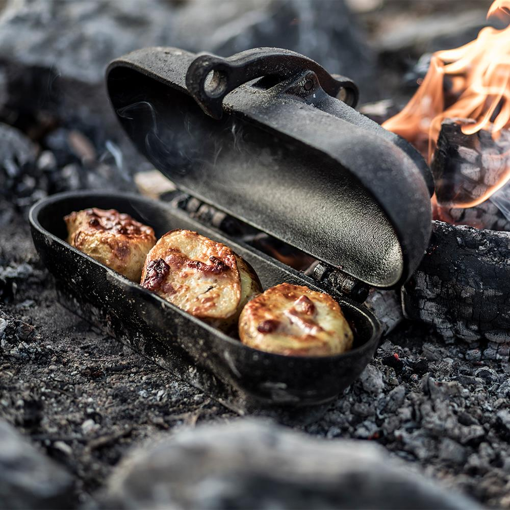 Gusseiserner Kartoffelbräter mit gebräunten Kartoffeln in Glut und Flammen