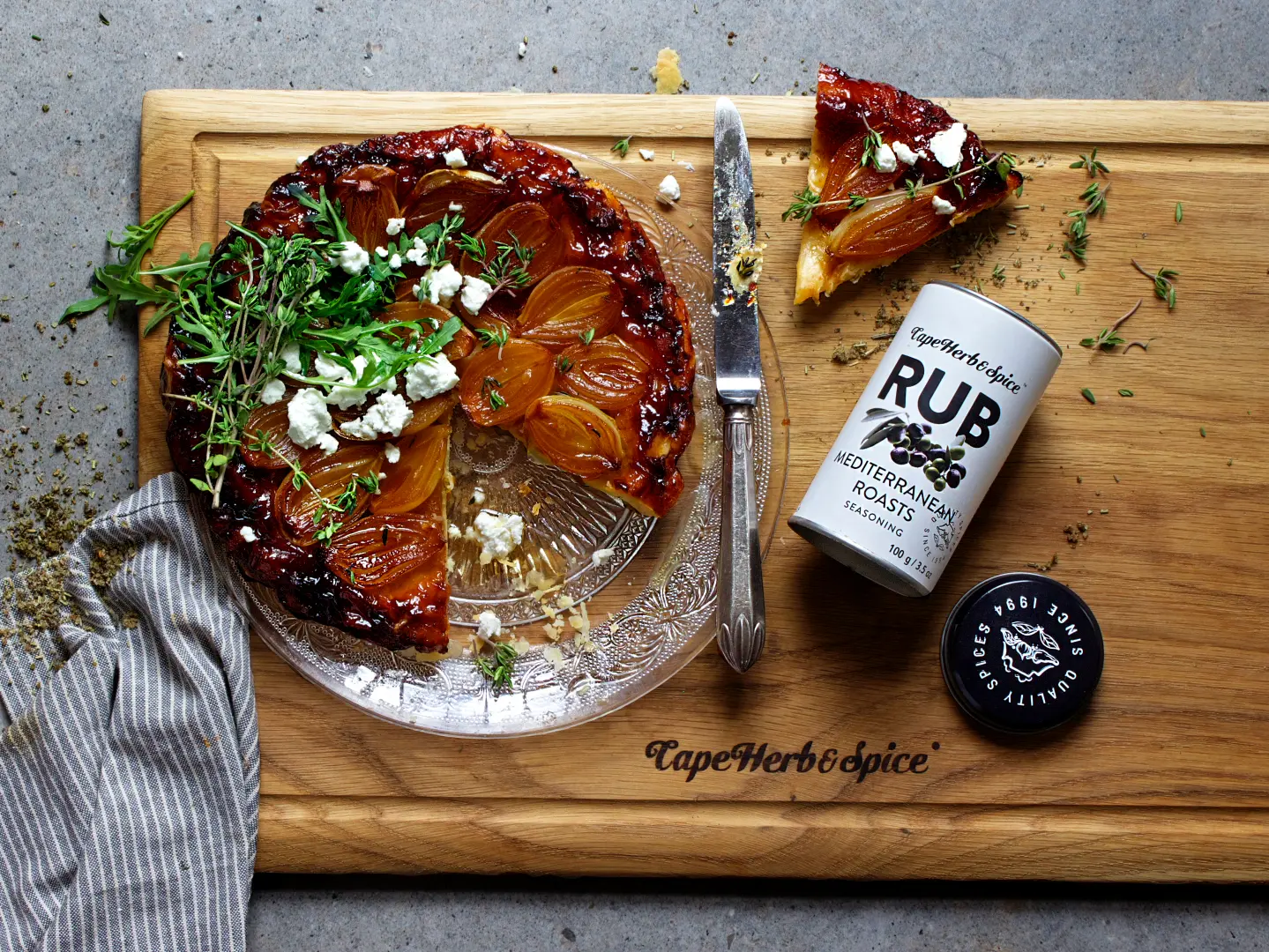 Tarte Tatin garniert mit Kräutern und Käse neben Cape Herb Rub Gewürzdose