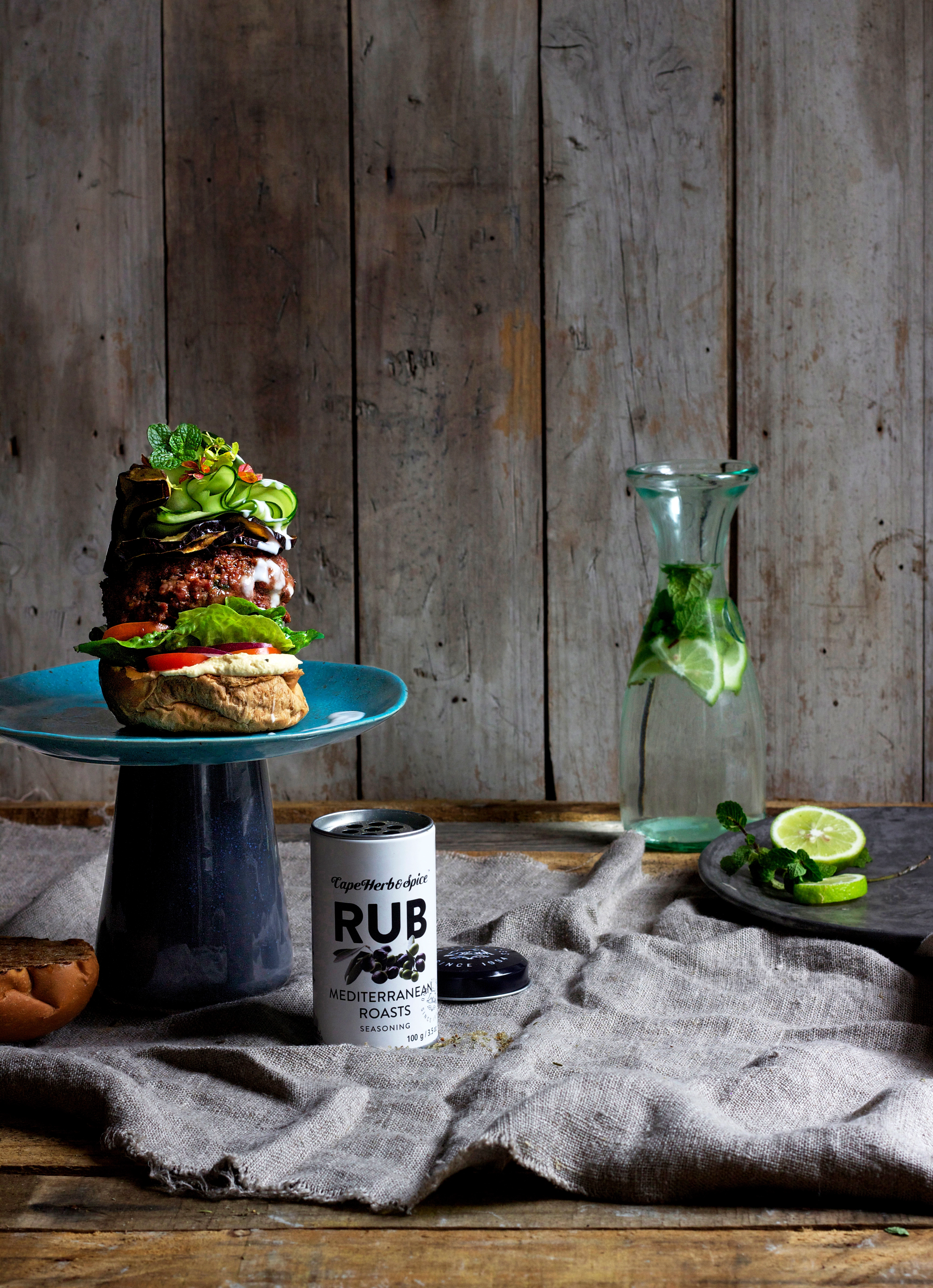 Rustikaler Burger auf blauem Teller mit Cape Herb Rub Gewürzdose und frischen Limetten