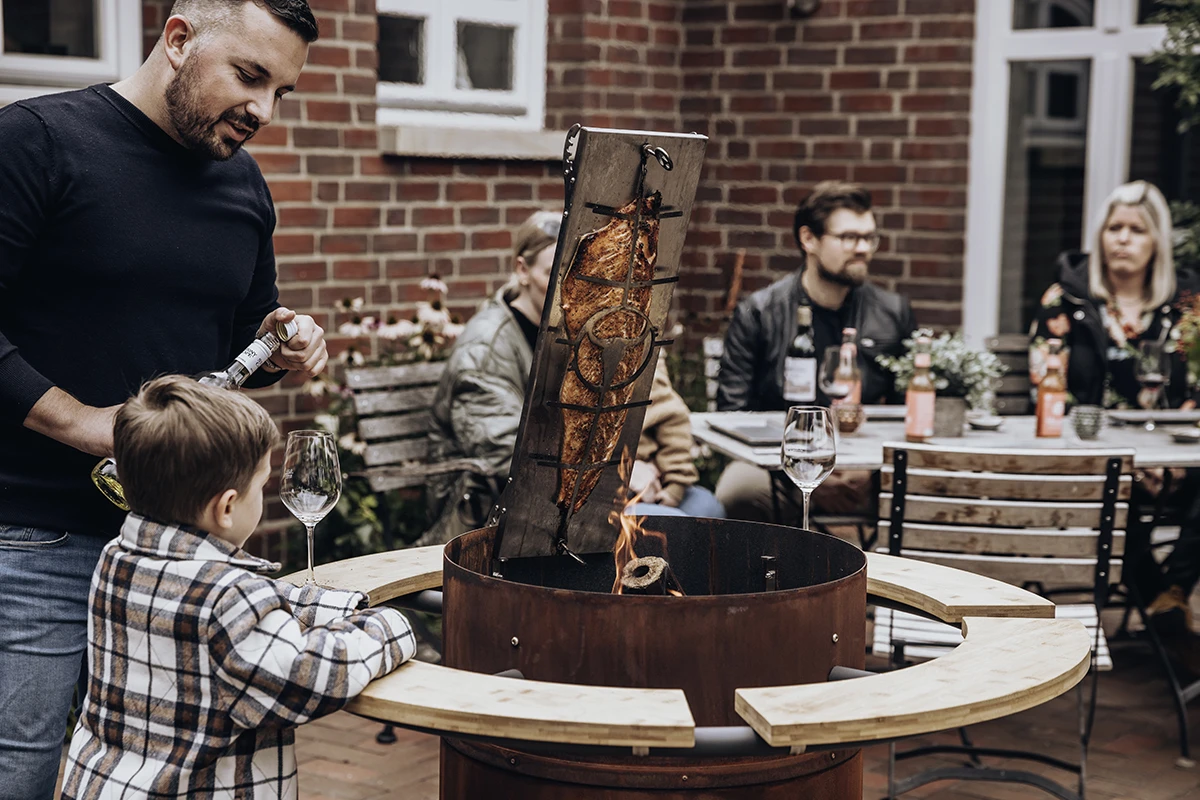 Mann und Junge grillen Fleisch am offenen Feuer im Garten, Gäste im Hintergrund
