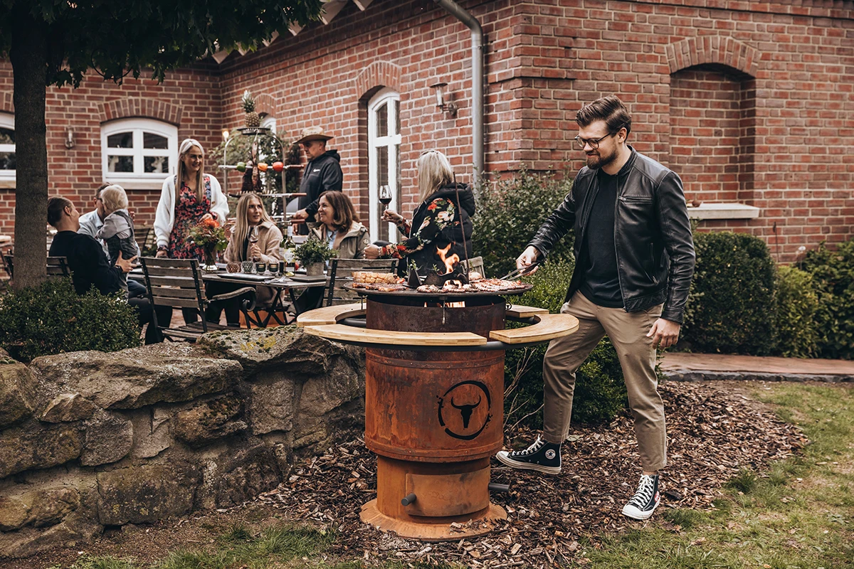 Mann grillt Pizza im Garten auf rustikalem Grill, umgeben von Gästen