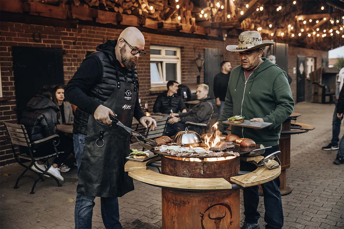 Zwei Männer grillen im Freien auf einem rustikalen Grill bei einer geselligen Zusammenkunft