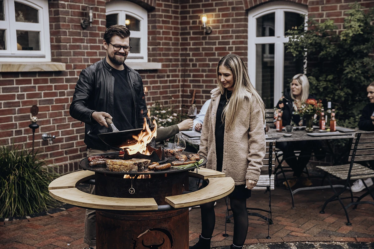 Mann grillt mit Flammen in Pfanne neben Frau im Garten bei geselligem Beisammensein