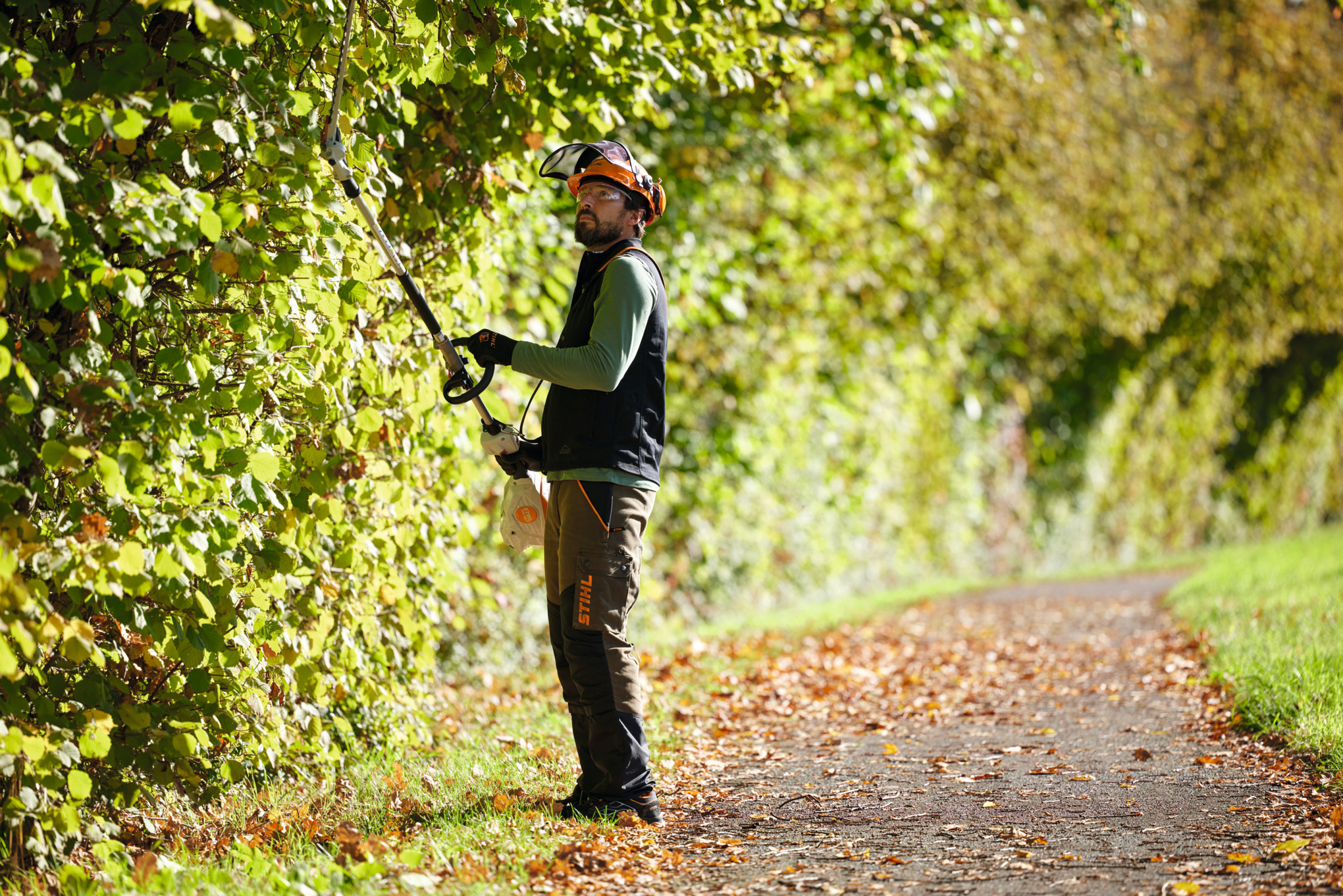 Mann verwendet Stihl KMA 120 R Akku-KombiMotor für Gartenarbeit auf einem sonnigen Waldweg