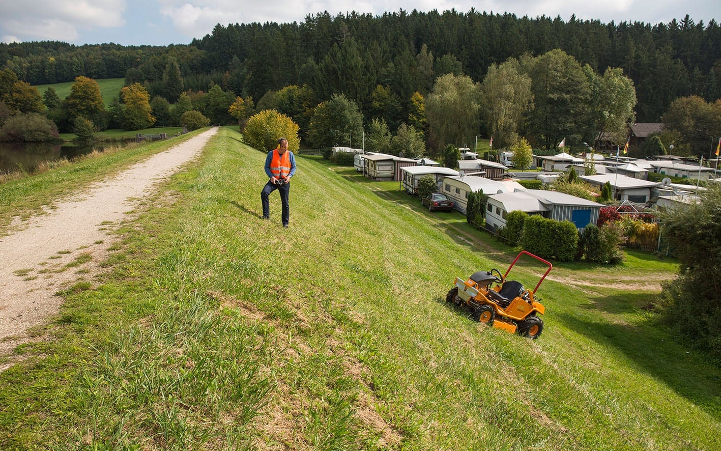 Mann bedient ferngesteuerten Aufsitzmäher AS 940 Sherpa auf Hang neben Campingplatz